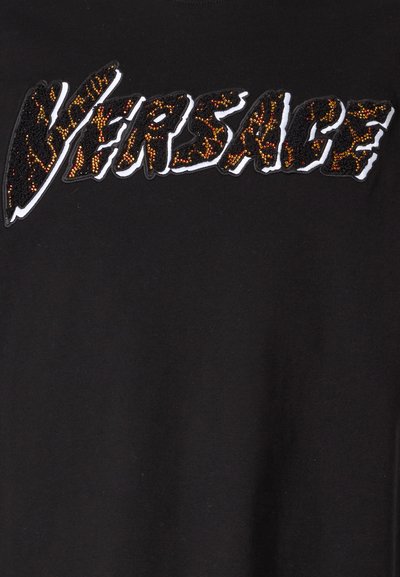 Melna T-kreklu ar izteiksmīgu, sekvīnu "VERSACE" logo šķeltnes fontā, ar zelta un brūnām akcentiem uz baltas kontūras.