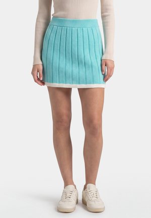 Woman wearing a short turquoise ribbed skirt with white trim, beige ribbed long-sleeve top, and white sneakers, standing against a plain background.