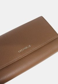 Portafoglio in pelle marrone con superficie texture, caratterizzato da un logo dorato "Coccinelle" impresso sulla parte anteriore. Design a chiusura con pattina, forma rettangolare.