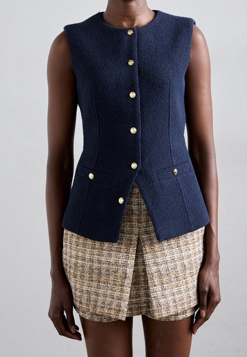 Veste sans manches bleu marine texturée avec des boutons dorés portée sur une jupe en tweed beige et marron avec une fente à l'avant.