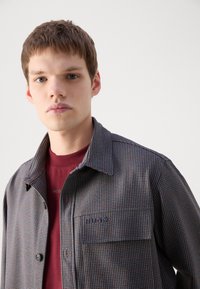 Checkered button-up jacket in gray, red, and blue fabric, featuring a chest pocket and subtle branding. Worn over a maroon shirt.