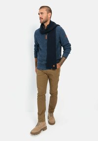 Blue knitted sweater with a ribbed navy scarf, tan chinos, and light brown boots. Notable details: leather tag on sweater and scarf texture.