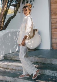 Witte oversized knoop-up blouse, haakbroek en chunky sandalen. Lichte textuur op de stof. Grote, gebogen handtas in een neutrale kleur.