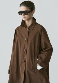 Woman with pulled-back hair wearing tortoiseshell sunglasses and an oversized brown buttoned coat, standing against a plain background.