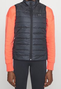 Musta puffertakki, jossa on vetoketju edessä ja vaakasuorat tikatut kuviot, yhdistettynä kirjavaan oranssiin pitkähihaiseen paitaiseen sen alla.