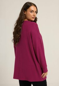 Pull à manches longues en fuchsia, coupe ample, tissu doux, encolure ronde, fentes latérales, texture lisse et poignets côtelés.