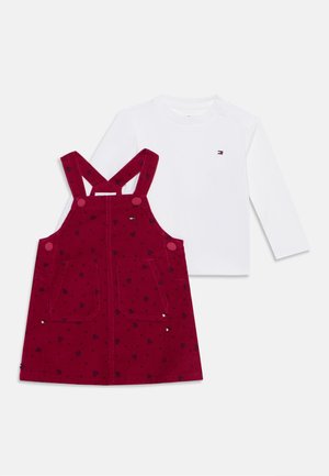 Chemise blanche à manches longues associée à une robe tablier en velours côtelé rouge avec des motifs de cœurs noirs et des bretelles boutonnées.