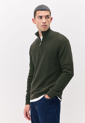 Young man wearing dark green textured zip-up sweater and blue jeans, looking to his left against a plain white background.