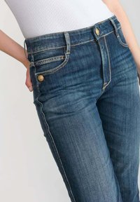 Jeans en denim bleu foncé au design ajusté, dotés de boutons dorés, de poches avant et de surpiqûres contrastées. Texture lisse et jambe droite.