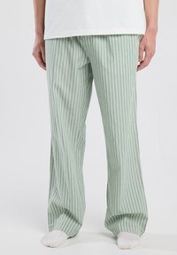 Persoon in groene en witte verticaal gestreepte losse broek met witte sokken, staande tegen een effen lichte achtergrond.