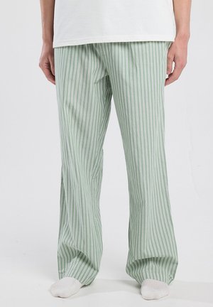 Persoon in groene en witte verticaal gestreepte losse broek met witte sokken, staande tegen een effen lichte achtergrond.