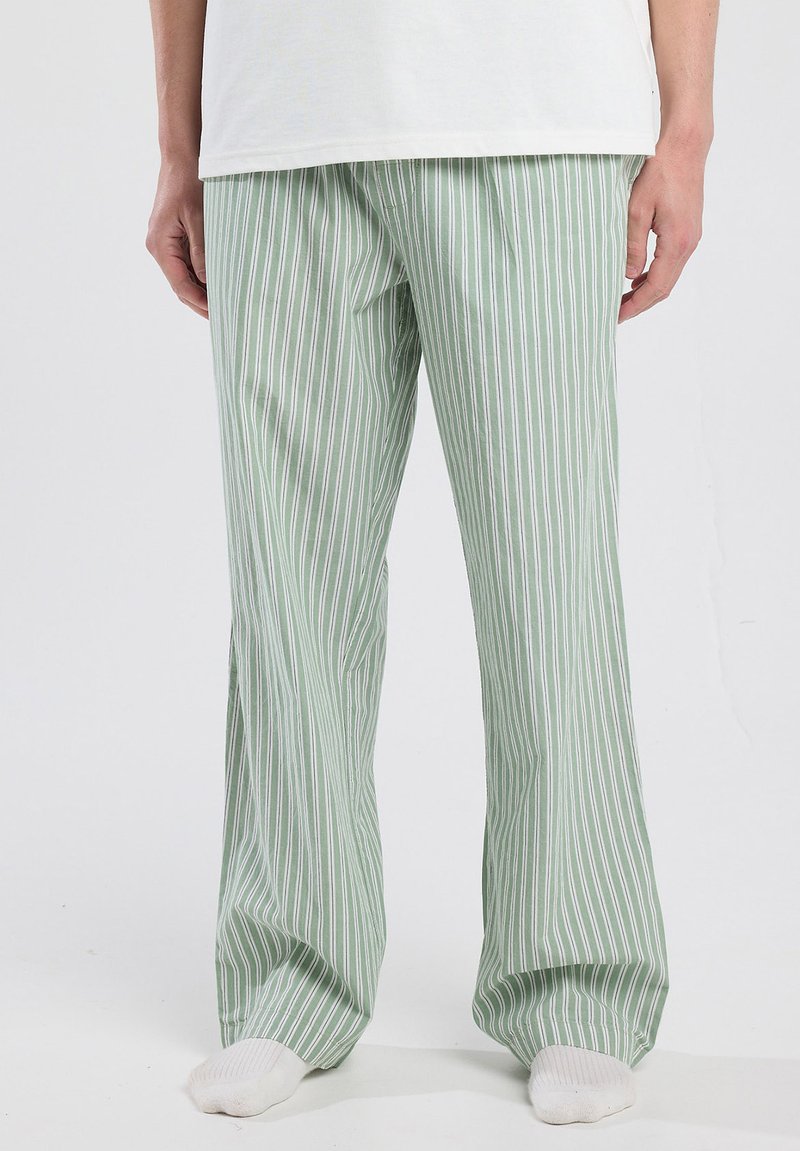 Persoon in groene en witte verticaal gestreepte losse broek met witte sokken, staande tegen een effen lichte achtergrond.
