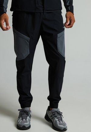 Hombre que lleva pantalones deportivos negros con parches grises en las rodillas y zapatillas deportivas grises, de pie frente a un fondo liso.
