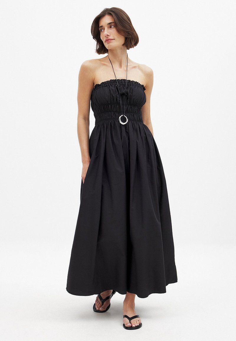Robe maxi noire sans bretelles avec un corsage froncé et une jupe ample ; présente une taille cintrée et un tissu lisse et léger. Tongs noires.