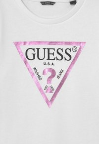 T-shirt in cotone bianco con un grande logo triangolare rosa e nero, accompagnato dal testo "GUESS U.S.A." e "WASHED JEANS" e un design a forma di punto interrogativo.