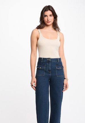 Femme portant un haut beige sans manches avec des bretelles tressées et un jean bleu marine taille haute avec des poches à rabat à l'avant.