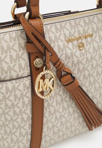 Beige Canvas-Handtasche mit Monogramm-Muster, braunen Lederakzenten, goldenen Beschlägen und einem Quasten-Detail mit einem runden Logo-Anhänger.