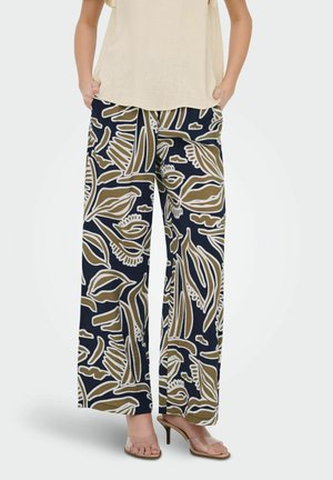 Mujer con pantalones de pierna ancha con patrones abstractos de hojas en azul marino, marrón y blanco, combinados con top beige y tacones con tiras transparentes.