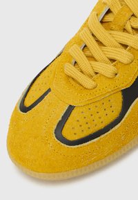 Gelbe Sneaker mit einer strukturierten Wildleder-Oberfläche, schwarzen Akzenten, durchlöcherten Details und flachen gelben Schnürsenkeln sowie einer transparenten Gummisohle.