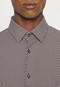 Gros plan d’une personne portant une chemise boutonnée à motifs géométriques marron, blanc et noir avec un col pointu.