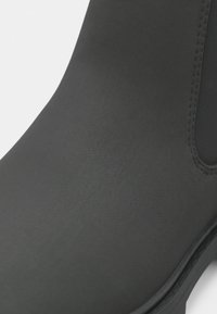 Gros plan d’une chaussure en cuir noir lisse avec des coutures visibles et un bout arrondi sur un fond clair uni.