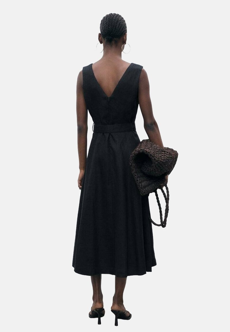 Femme aux cheveux tressés portant une robe midi noire sans manches et des talons noirs, tenant un grand sac tressé foncé, dos à la caméra.