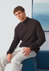 Donkerbruin pullover trui met een hoge kraag en rits, met een geëtst diamantenpatroon. Gepaard met lichtgekleurde broek.