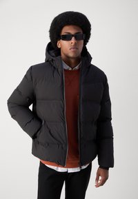 Czarna kurtka puchowa z kapturem, z zamkiem z przodu, grubą ocieplaną konstrukcją oraz elastycznymi mankietami. Noszona na pomarańczowym swetrze.