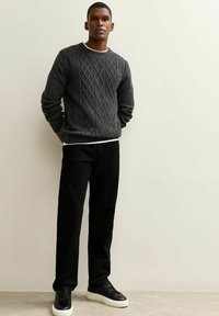 Jersey de punto grueso gris oscuro con cuello redondo, combinado con pantalones negros y zapatillas negras con suelas blancas, de pie contra un fondo neutral.