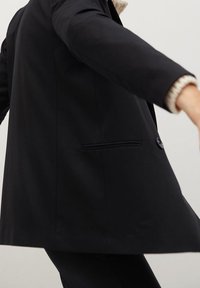 Personne portant un blazer noir par-dessus un pull crème, en mouvement sur un fond clair uni.