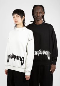 Deux modèles portant des pulls surdimensionnés. L'un est blanc avec du texte noir en bas ; l'autre est noir avec du texte blanc. Les deux ont des poignets côtelés.