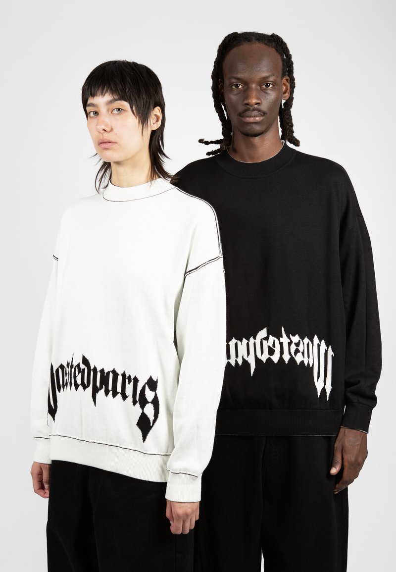 Deux modèles portant des pulls surdimensionnés. L'un est blanc avec du texte noir en bas ; l'autre est noir avec du texte blanc. Les deux ont des poignets côtelés.