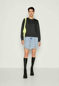 Mulher vestindo um sweatshirt preto, saia de ganga azul clara com fendas frontais, botas pretas até o joelho e uma mala de ombro amarelo-neon está encostada a um fundo simples.