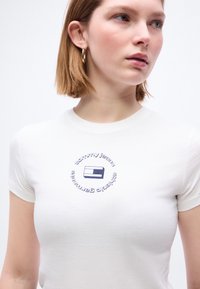 Νεαρή γυναίκα με κοντά ανοιχτό καστανά μαλλιά, που φοράει λευκό μπλουζάκι με κυκλικό λογότυπο Tommy Jeans και χρυσό κρίκο στο αυτί, κοιτάζει δεξιά.