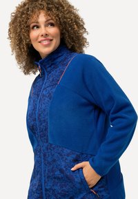 Veste en polaire bleue avec une partie supérieure à motifs, fermeture éclair sur le devant et accents orange. Comprend des poches sur les côtés avant.