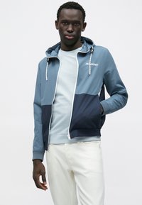 Ανδρικό μπουφάν με κουκούλα σε ανοιχτό μπλε και ναυτικό χρώμα, κατασκευασμένο από λείο υλικό, με φερμουάρ μπροστά, ελαστικές μανσέτες και λεπτομέρεια με λογότυπο.
