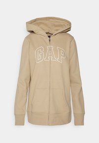 Sudadera con capucha color beige con cremallera, con un gran logo blanco "GAP" en el pecho, que cuenta con capucha y dos bolsillos frontales. Tela suave con puños acanalados.