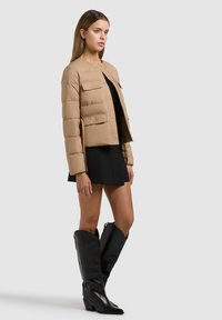 Žena v béžové prošívané bundě přes černé šaty s černými koženými kozačkami nad kolena, stojící před jednobarevným světlým pozadím.
