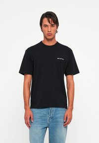 Mann iført svart Belstaff t-skjorte og lyseblå jeans, som står med ansiktet vendt fremover mot en ensfarget hvit bakgrunn.