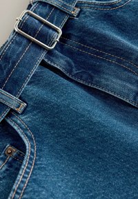 Spijkerbroek-overalls met een zilveren metalen gesp, donkerblauwe kleur, contrasterende oranje stiksels en een gladde textuur. Voorzien van een voorzak.