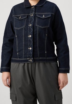 Mørkeblå denimjakke med hvide kontrastsømme og knapforlommen på brystet, båret over mørke cargobukser.