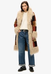 Abrigo largo de piel sintética con diseño de patchwork multicolor, que presenta un amplio collar y puños peludos, combinado con unos vaqueros azules remangados y zapatos negros.