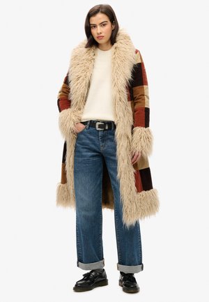 Faux fur long coat with multicolored patchwork design, featuring a wide furry collar and cuffs, paired with blue rolled-up jeans and black shoes.