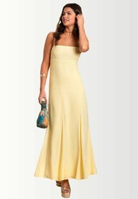 Gele maxi-jurk met een vierkante halslijn, een vloeiende silhouet en zijplooien. Model houdt een multikleurige tas vast en draagt plateauschoenen.