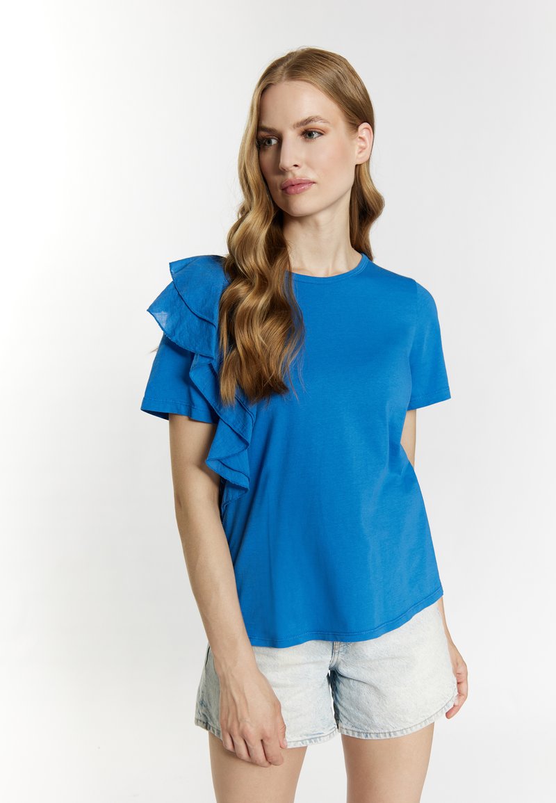 Blaues T-Shirt mit Rüschendetails an den geschichteten Ärmeln. Aus leichtem Stoff gefertigt, mit Rundhalsausschnitt und lässiger Passform. Kombiniert mit Jeansshorts.