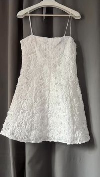 Witte jurk met decoratieve kantstructuur, mouwloos ontwerp, A-lijn vorm en een aansluitende lijfje. Hangt aan een lichtgekleurde hanger tegen een grijze achtergrond.