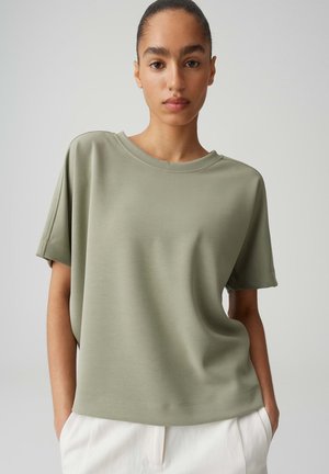 Vrouw met het haar naar achteren gebonden draagt losse lichtgroene korte mouw top en witte broek, staand tegen een effen lichte achtergrond.