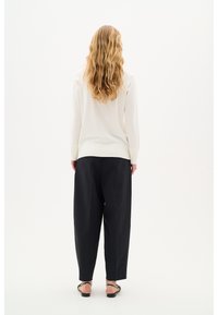 Weißer, gestrickter Pullover mit geripptem Saum, kombiniert mit lockeren, hochgeschnittenen schwarzen Hosen. Die Accessoires umfassen schwarze Sandalen. Schlichte Texturen.