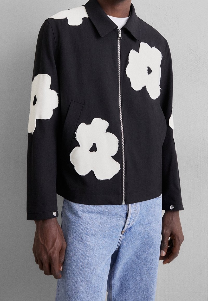 Chaqueta negra con cremallera que presenta grandes apliques florales blancos. Fabricada en tela, con mangas largas y collar. Detalle de bolsillo frontal visible.