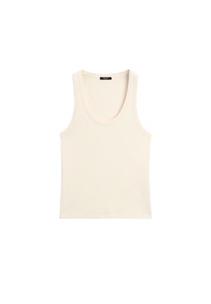 Frau geht drinnen, trägt ein beiges Tanktop, weiße weit geschnittene Hosen, schwarze Sandalen, Sonnenbrille und hält eine schwarze, strukturierte Tasche.